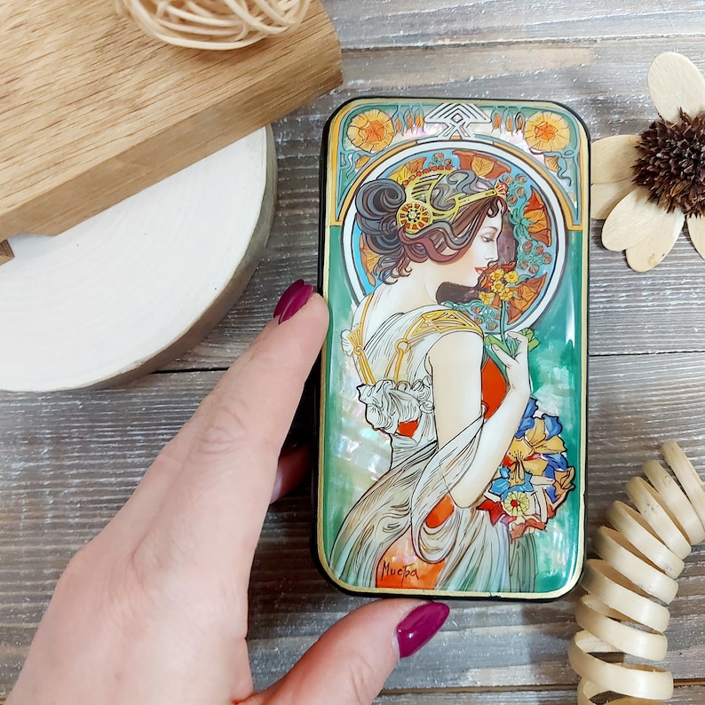 May include: A rectangular wooden box with a hinged lid. The lid features a colorful illustration of a woman in a white dress holding a bouquet of flowers. The illustration is in the Art Nouveau style. The box is signed "Mucha" in the lower right corner.