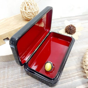 May include: A small black lacquered wooden box with a red interior. The box is open and a ring with a yellow stone is inside.
