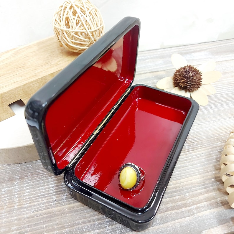 May include: A small black lacquered wooden box with a red interior. The box is open and a ring with a yellow stone is inside.