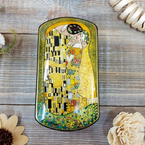 May include: Rectangular decorative box featuring Gustav Klimt's "The Kiss" painting. The box has a black border and a glossy finish. The artwork depicts a couple embracing, with gold, yellow, and black geometric patterns.