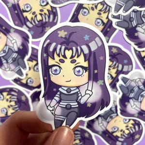 May include: A sticker featuring a cartoon character with purple hair, a white and black striped outfit, and a third eye. The character has a light pink blush and is surrounded by small stars and flowers.