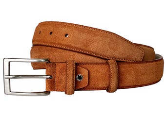 May include: A tan suede belt featuring a rectangular silver-toned metal buckle and a matching leather keeper loop. The belt strap has visible stitching along the edges and a small circular metal stud detail near the buckle with engraved text.