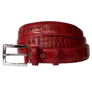 May include: A bright red belt with a glossy, embossed crocodile skin texture. The belt features a polished silver-tone metal rectangular buckle, a matching red leather keeper loop, and a small silver-tone metal stud detail near the buckle.