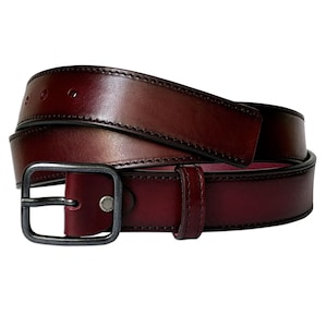 Peut inclure: Une ceinture en cuir bordeaux profond dotée d'une boucle rectangulaire en métal foncé et d'un passant en cuir assorti. La sangle présente des coutures visibles sur les bords et un petit rivet métallique près de la boucle marqué Made in Spain.