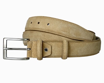 May include: A tan suede belt featuring a rectangular silver-toned metal buckle, two matching belt loops, and a small circular metal rivet detail on the strap near the buckle. The belt has visible stitching along the edges and multiple adjustment holes.