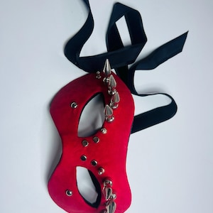Puede incluir: Una máscara de mascarada roja con púas y tachuelas plateadas. La máscara tiene una cinta negra para atar. La máscara tiene agujeros para los ojos y una forma curva, adecuada para un disfraz o evento temático.