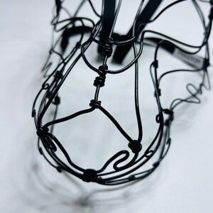 May include: A black wire sculpture of a horse's head, intricately crafted. The sculpture is made from thin, intertwined wires, forming a three-dimensional shape. The horse's head is set against a white background, emphasising the dark silhouette.