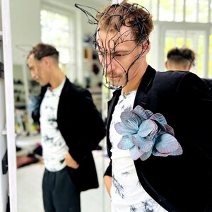 May include: A black blazer with a large, light blue and purple flower pinned to the lapel. The person is wearing a white t-shirt with a black tie-dye pattern and a black wire mask.