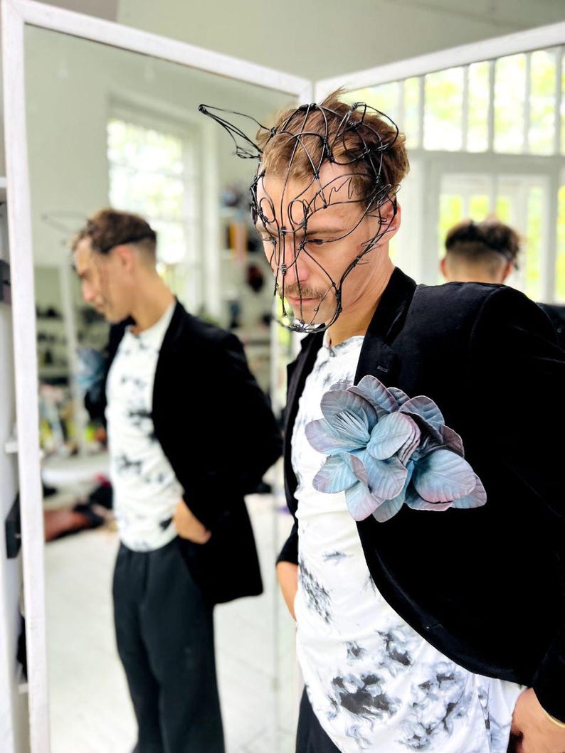 May include: A black blazer with a large, light blue and purple flower pinned to the lapel. The person is wearing a white t-shirt with a black tie-dye pattern and a black wire mask.