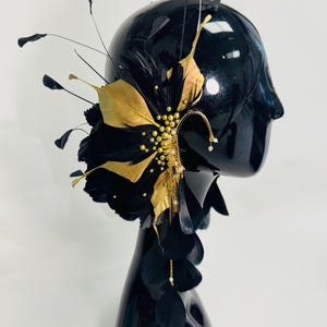 May include: An ear cuff featuring black and gold feathers, accented with small gold beads and delicate gold chains. The piece is designed to wrap around the ear, with long black feathers extending outward and downward, creating a layered, textured appearance.