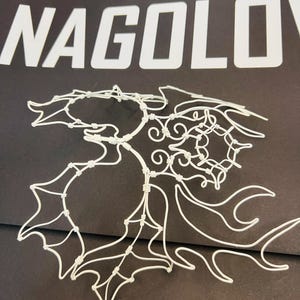 May include: A white wire sculpture in the shape of a leaf, with intricate details and a delicate appearance. The sculpture is set against a dark background, with the word "NAGOLOV" in white block letters above it.