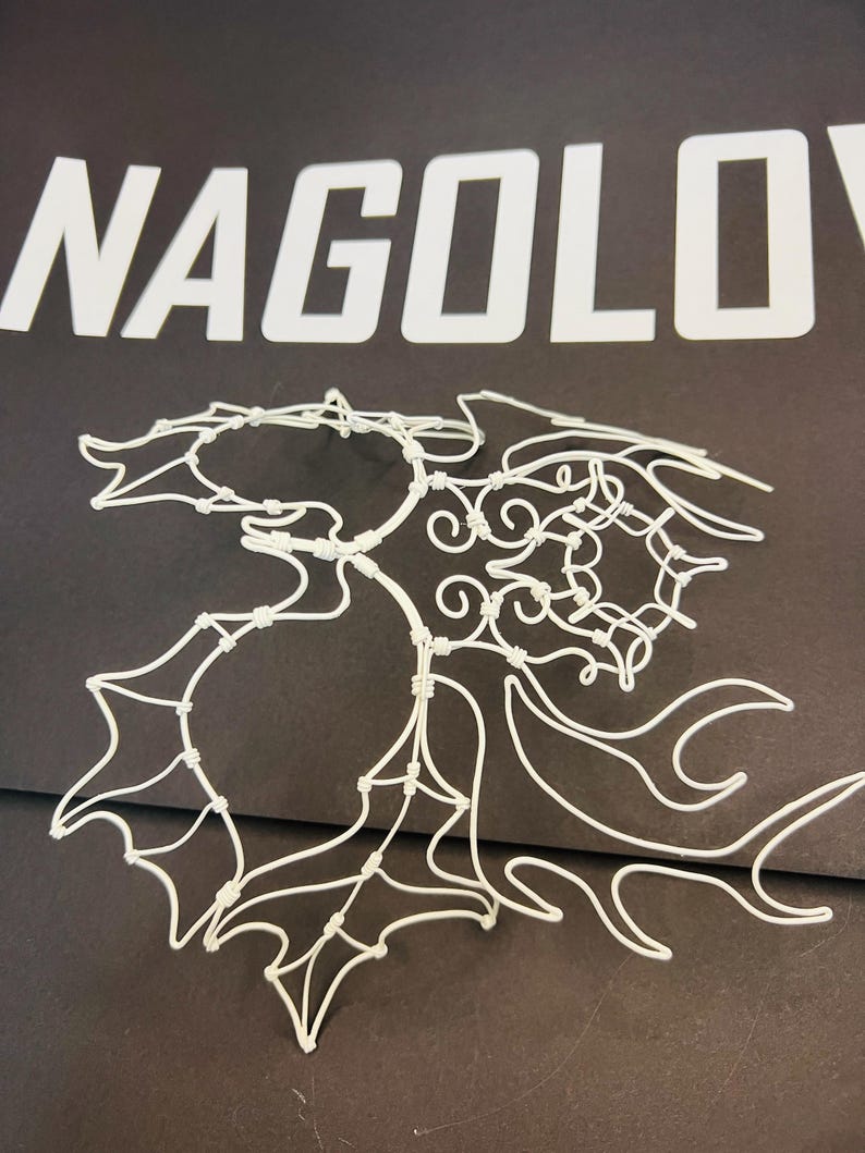 May include: A white wire sculpture in the shape of a leaf, with intricate details and a delicate appearance. The sculpture is set against a dark background, with the word "NAGOLOV" in white block letters above it.
