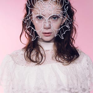 May include: A white wire mask with an intricate design, worn over the eyes and forehead. The mask is the focal point, set against a pink background. The person is wearing a white lace top. The overall aesthetic is ethereal and artistic.
