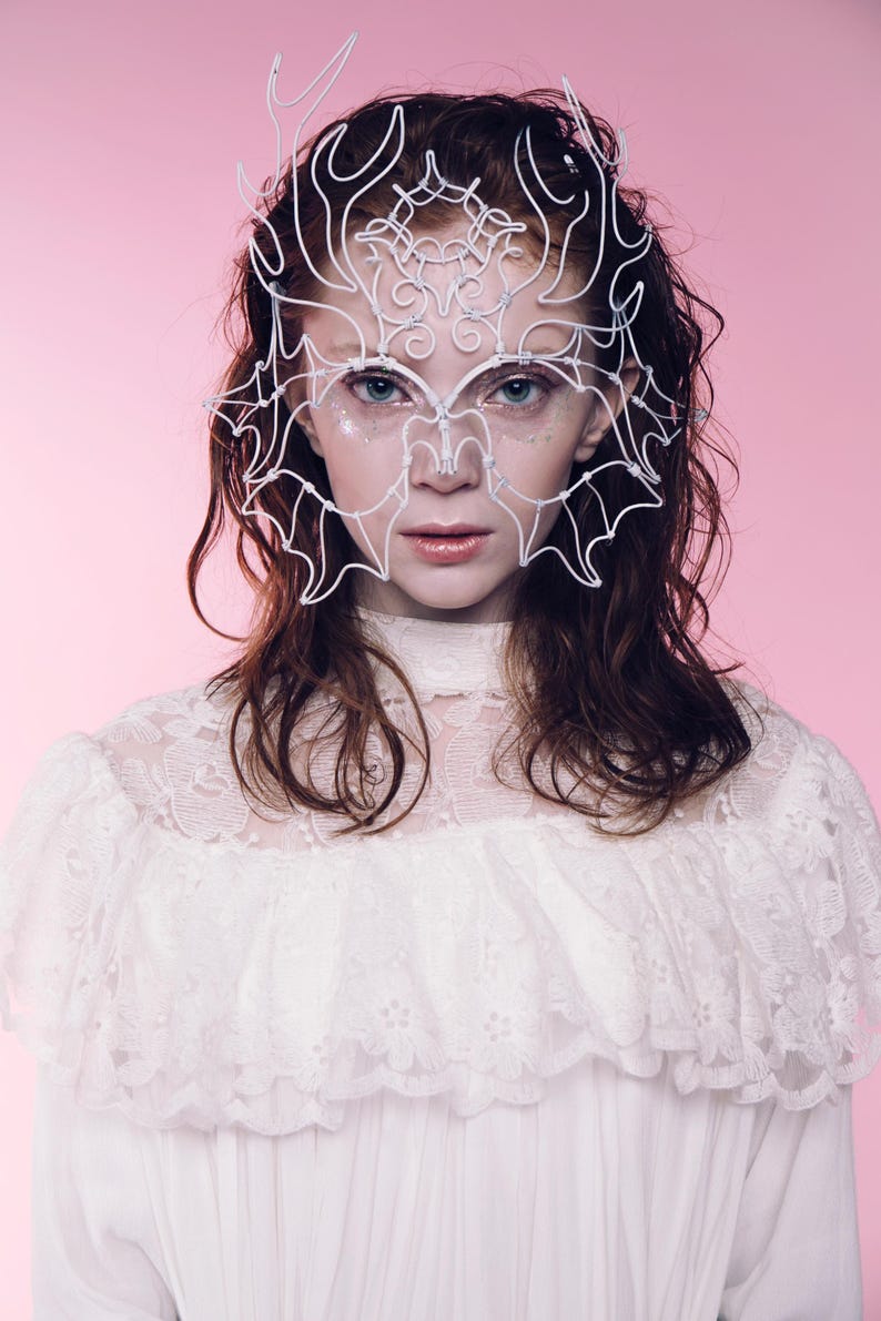May include: A white wire mask with an intricate design, worn over the eyes and forehead. The mask is the focal point, set against a pink background. The person is wearing a white lace top. The overall aesthetic is ethereal and artistic.