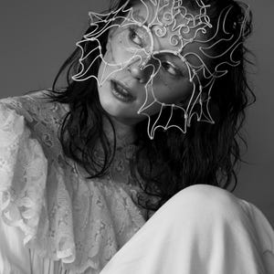 May include: Black and white portrait of a person wearing a delicate, wire-frame mask. The mask covers the upper face, with an intricate, abstract design. The person is wearing a white lace top and a white dress.