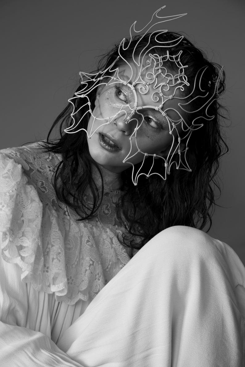 May include: Black and white portrait of a person wearing a delicate, wire-frame mask. The mask covers the upper face, with an intricate, abstract design. The person is wearing a white lace top and a white dress.