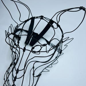 May include: A black wire fox head sculpture with long ears and a detailed facial structure. The mask has a black strap for wearing and is set against a white background. The sculpture is made of thin, intertwined wires.