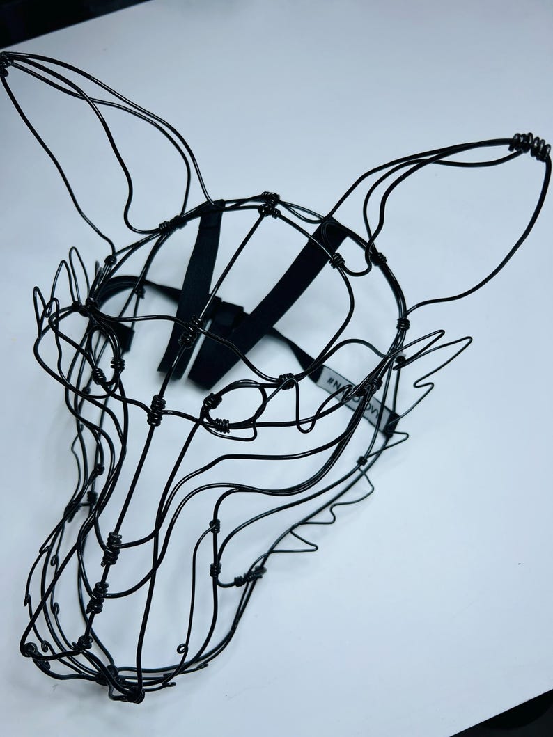 May include: A black wire fox head sculpture with long ears and a detailed facial structure. The mask has a black strap for wearing and is set against a white background. The sculpture is made of thin, intertwined wires.