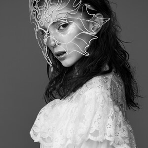 May include: Black and white portrait of a person wearing a delicate, white wire mask with an intricate, nature-inspired design. The person is also wearing a white lace top. The background is a solid gray.