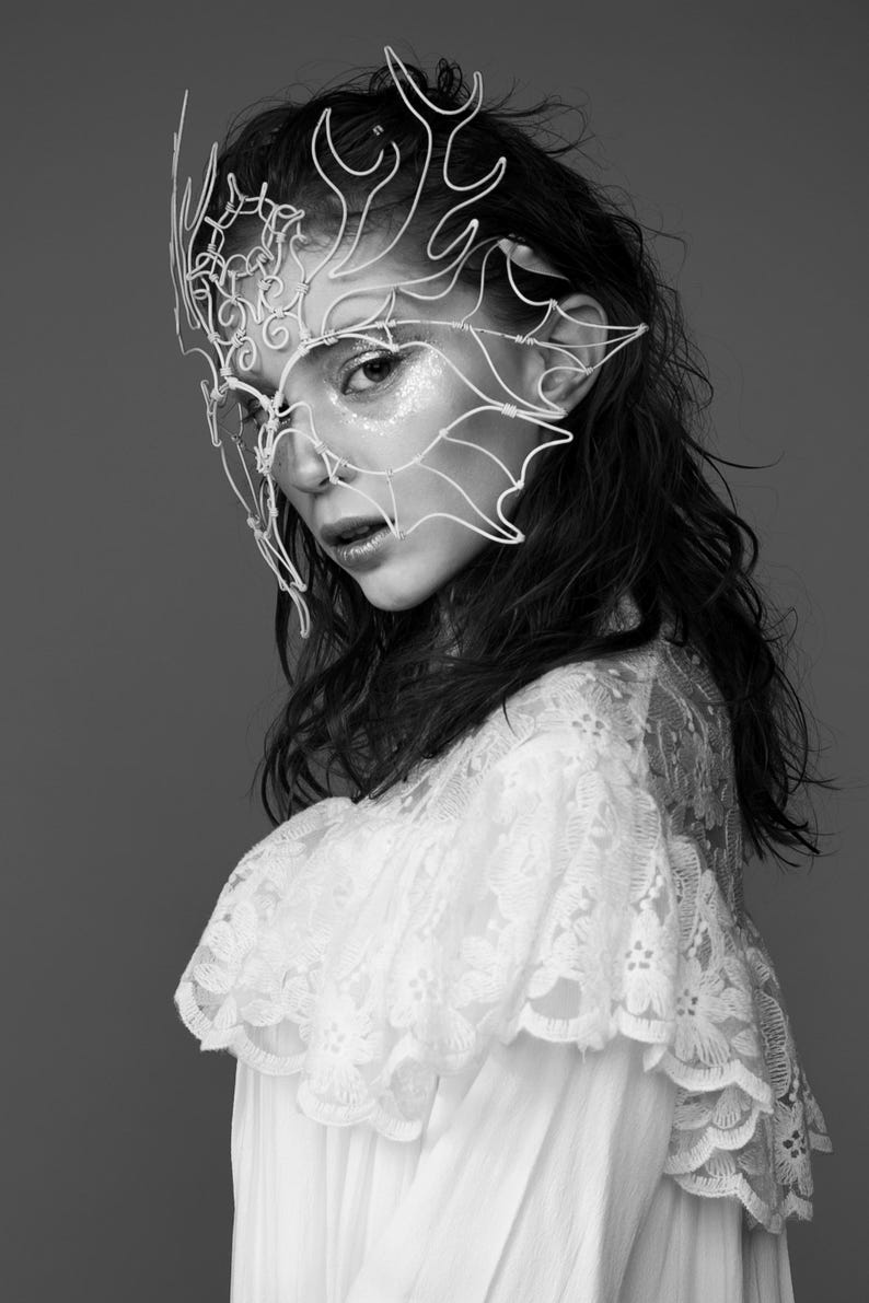 May include: Black and white portrait of a person wearing a delicate, white wire mask with an intricate, nature-inspired design. The person is also wearing a white lace top. The background is a solid gray.