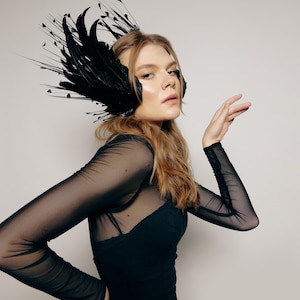 Può includere: Un copricapo nero di piume dal design drammatico. Il copricapo presenta grandi piume curve e delicati accenti a punta. La modella indossa un abito nero con maniche trasparenti.