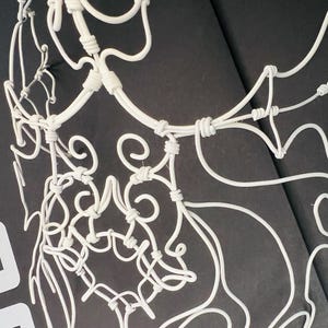 May include: A white, wire-sculpted decorative piece with intricate, swirling designs. The artwork features a network of curved lines and knots, creating a delicate, openwork pattern. The piece is set against a dark background, highlighting its form.