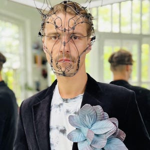 May include: A person wearing a wire sculpture mask resembling a rabbit's face. The mask is crafted from thin, dark wire. They are also wearing a black velvet blazer, adorned with a large, blue and purple flower pinned to the lapel.