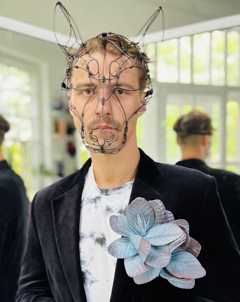 May include: A person wearing a wire sculpture mask resembling a rabbit's face. The mask is crafted from thin, dark wire. They are also wearing a black velvet blazer, adorned with a large, blue and purple flower pinned to the lapel.