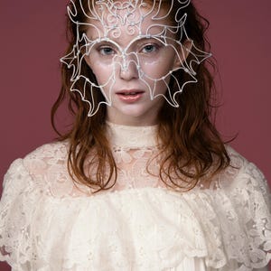 May include: A white wire mask with an intricate design, featuring a crown-like structure and bat-wing shapes, worn over the face. The person is wearing a white lace dress. The background is a solid maroon color.