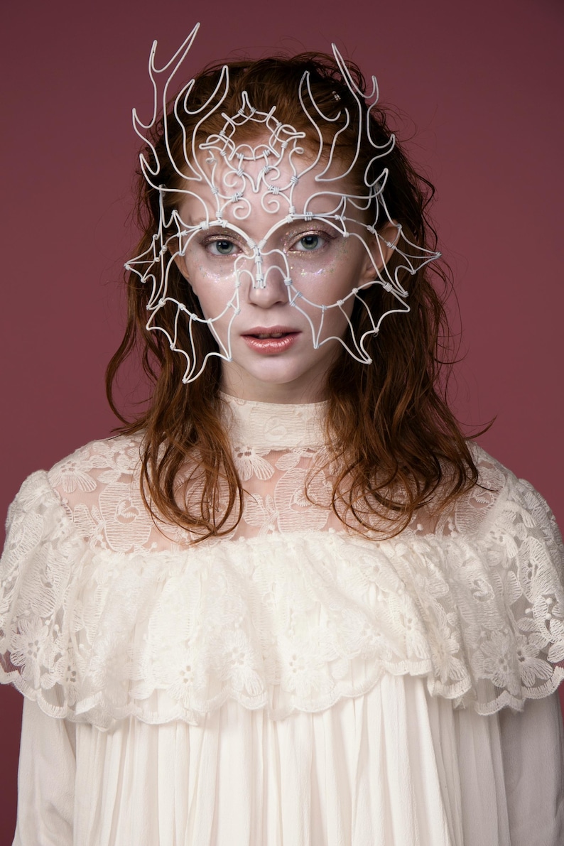 May include: A white wire mask with an intricate design, featuring a crown-like structure and bat-wing shapes, worn over the face. The person is wearing a white lace dress. The background is a solid maroon color.