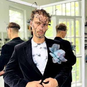May include: A black velvet blazer with a tie-dye t-shirt and a large, blue and grey flower pinned to the lapel. The person is wearing a wire mask. The background includes a mirror and a window.