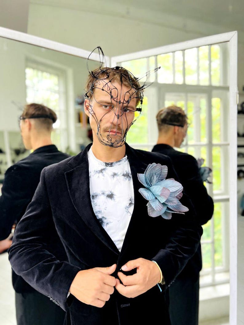 May include: A black velvet blazer with a tie-dye t-shirt and a large, blue and grey flower pinned to the lapel. The person is wearing a wire mask. The background includes a mirror and a window.
