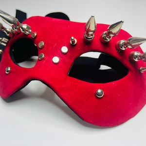 May include: A red masquerade mask with silver spikes and studs. The mask has eye cutouts and a black ribbon tie. The mask is made of a soft, velvety material.