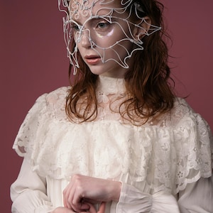 May include: A white wire mask with an intricate design, worn over the face. The mask is the focal point, set against a pink background. The person is wearing a white lace dress with a high collar and ruffled shoulders.