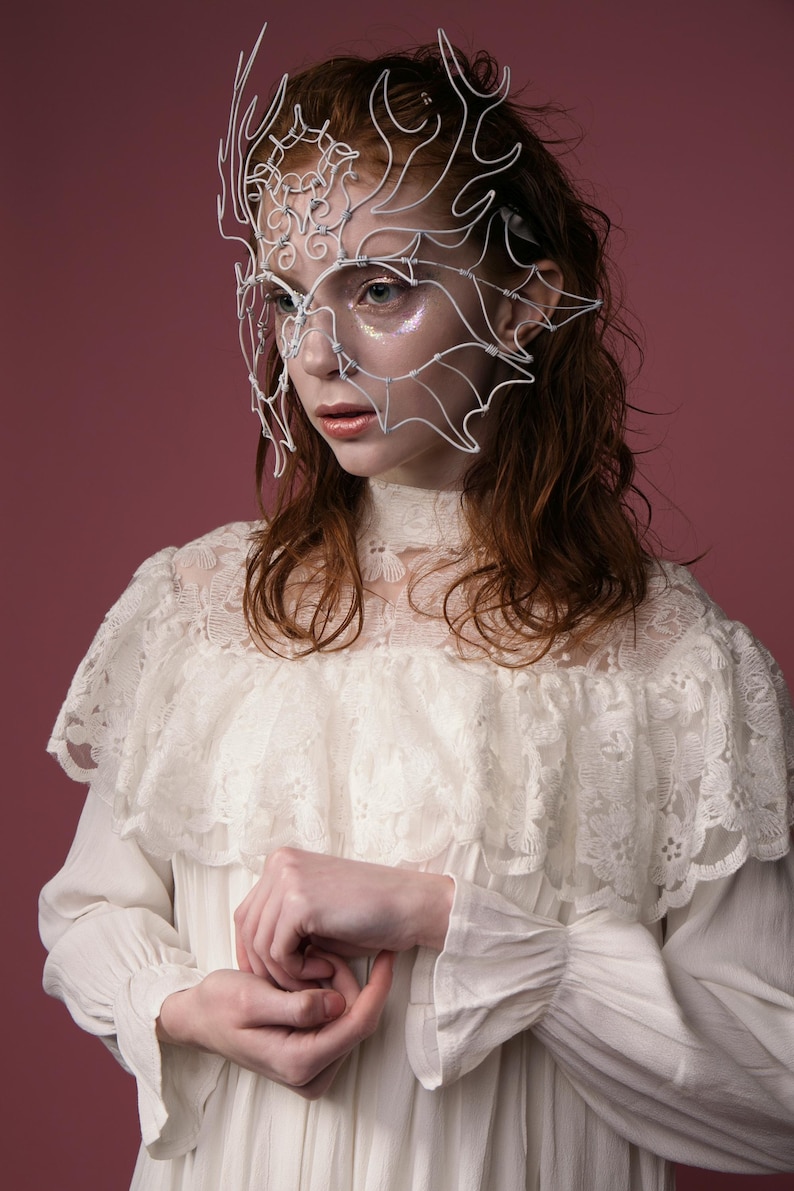 May include: A white wire mask with an intricate design, worn over the face. The mask is the focal point, set against a pink background. The person is wearing a white lace dress with a high collar and ruffled shoulders.
