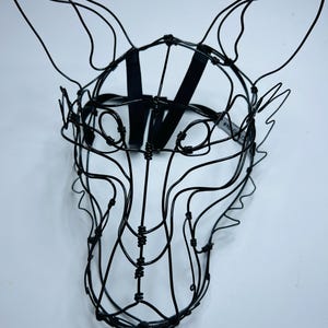 May include: A black wire sculpture of an animal head, possibly a fox or wolf, with large ears and a muzzle. The sculpture is made of thin, black wire and features a black strap for wearing. The design is intricate and artistic.