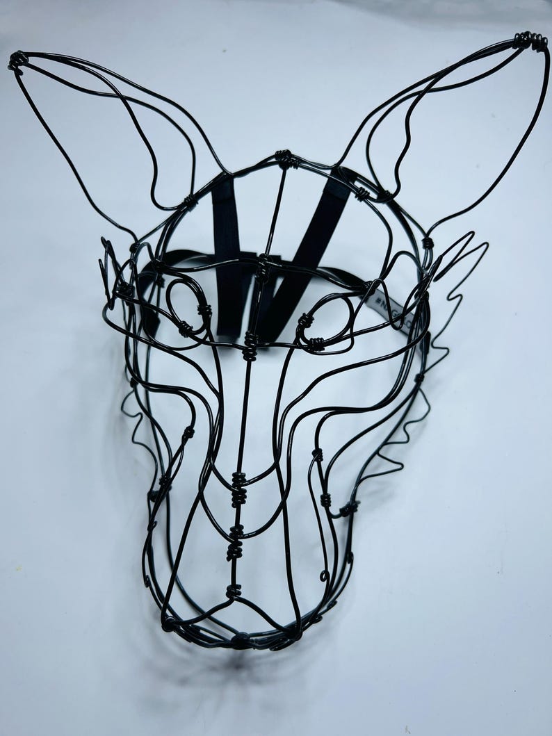 May include: A black wire sculpture of an animal head, possibly a fox or wolf, with large ears and a muzzle. The sculpture is made of thin, black wire and features a black strap for wearing. The design is intricate and artistic.