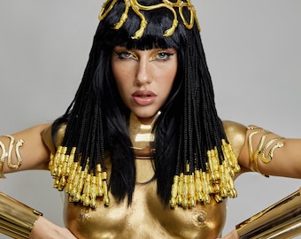 May include: A woman in a gold costume, reminiscent of ancient Egyptian royalty. The costume includes a gold headpiece, arm cuffs, and body paint. The black wig is styled with gold accents.