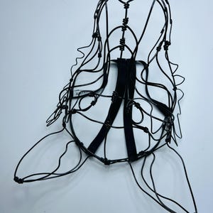 May include: A black wire sculpture of a head and shoulders, with a cage-like structure. The sculpture has a rounded top, flowing lines suggesting hair, and a flat base. Two black straps are attached to the back.