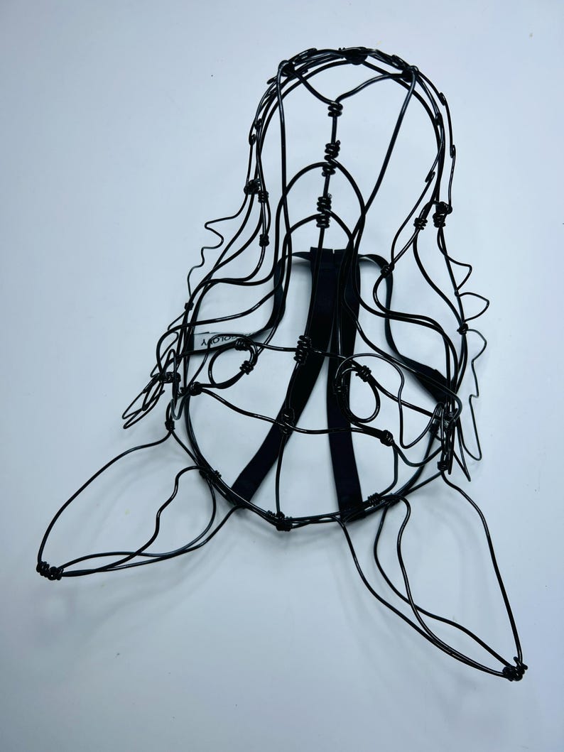 May include: A black wire sculpture of a head and shoulders, with a cage-like structure. The sculpture has a rounded top, flowing lines suggesting hair, and a flat base. Two black straps are attached to the back.