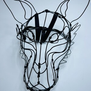 May include: A black wire fox mask sculpture. The mask is made from thin, black wire, formed into a fox's head with pointed ears. A black strap is attached for wearing. The mask is set against a white background.
