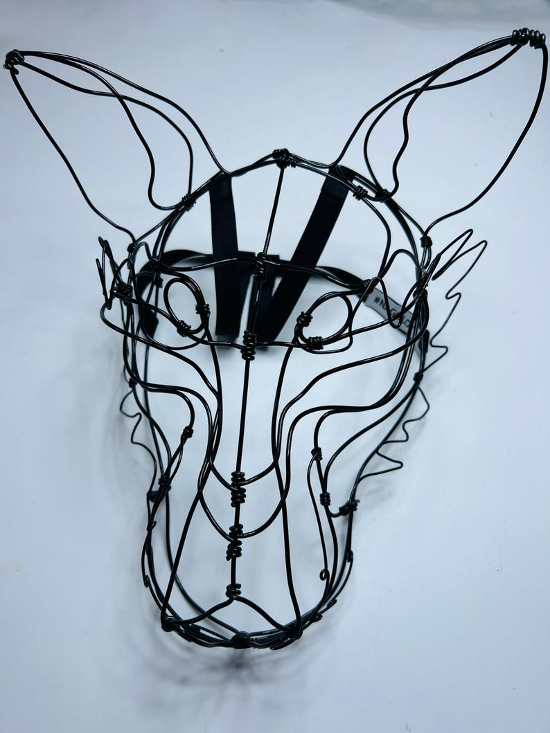 May include: A black wire fox mask sculpture. The mask is made from thin, black wire, formed into a fox's head with pointed ears. A black strap is attached for wearing. The mask is set against a white background.