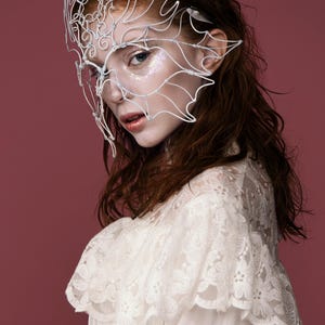 May include: A white wire mask with an intricate design, worn by a person with auburn hair and a white lace top. The mask covers the eyes and extends to the ears. The background is a solid maroon color.