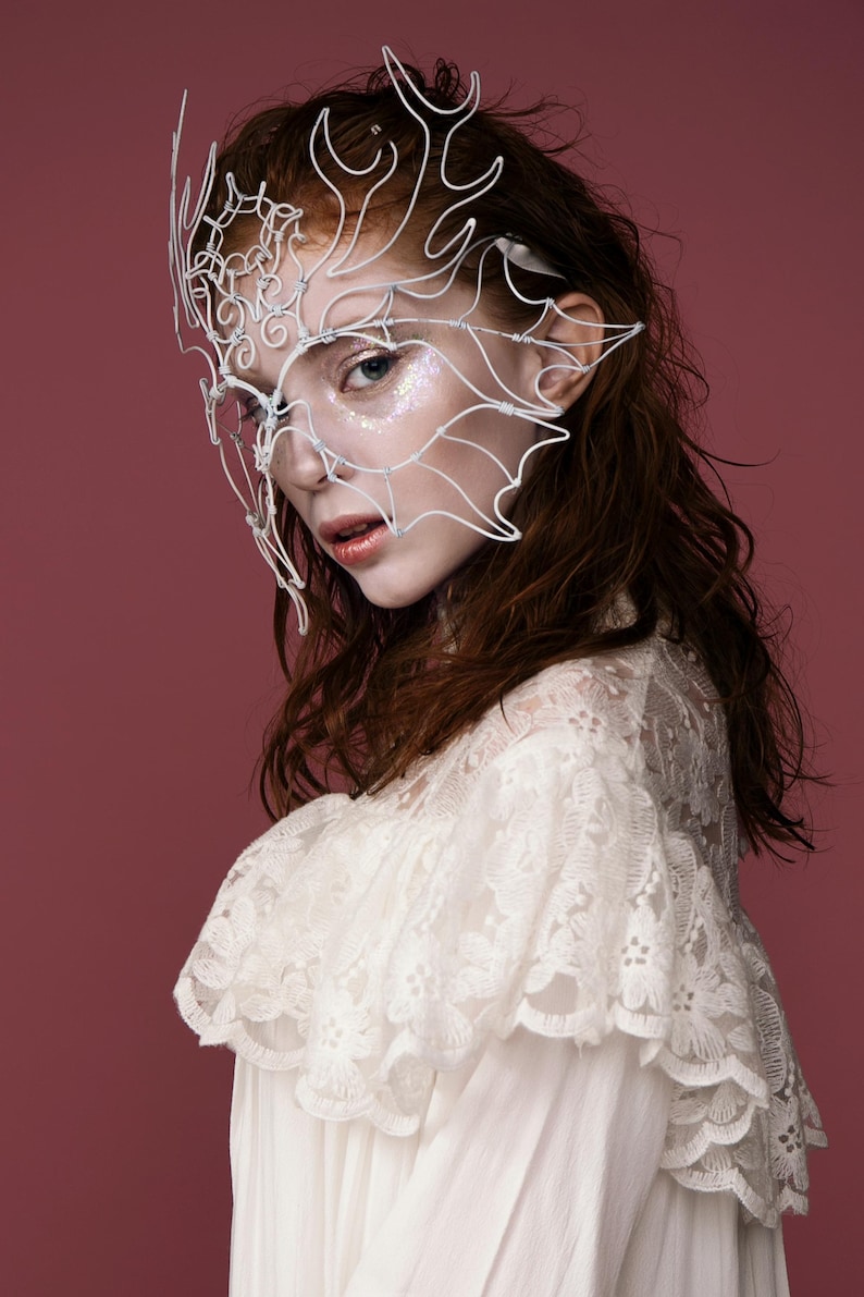 May include: A white wire mask with an intricate design, worn by a person with auburn hair and a white lace top. The mask covers the eyes and extends to the ears. The background is a solid maroon color.