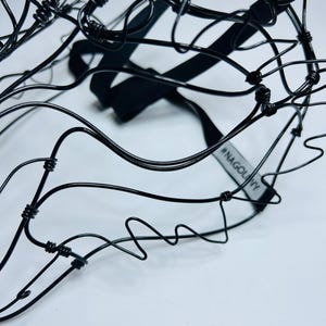May include: A close-up shot of a black wire sculpture with a complex, abstract form. The sculpture features a black elastic strap and a tag with the text "#NAGOLNY". The background is plain white.