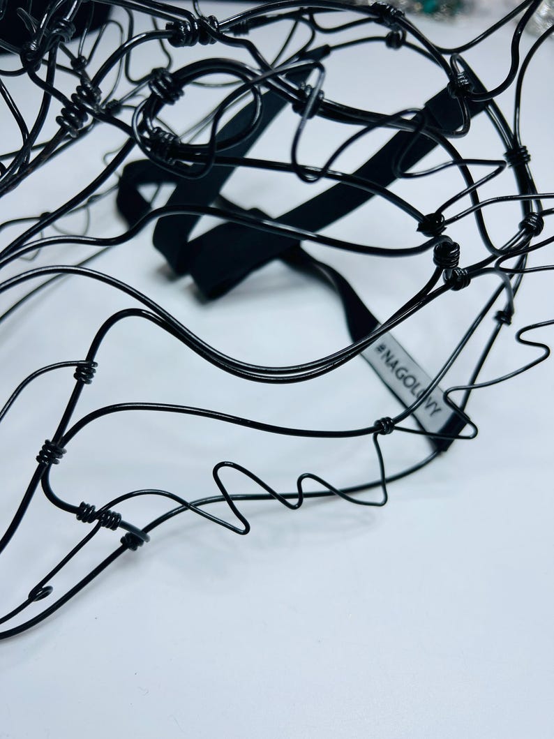 May include: A close-up shot of a black wire sculpture with a complex, abstract form. The sculpture features a black elastic strap and a tag with the text "#NAGOLNY". The background is plain white.