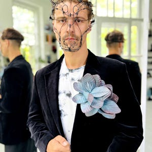 May include: A person wearing a black velvet blazer over a tie-dye t-shirt, adorned with a large, blue and purple flower. A wire sculpture mask with bunny ears is worn on the head.