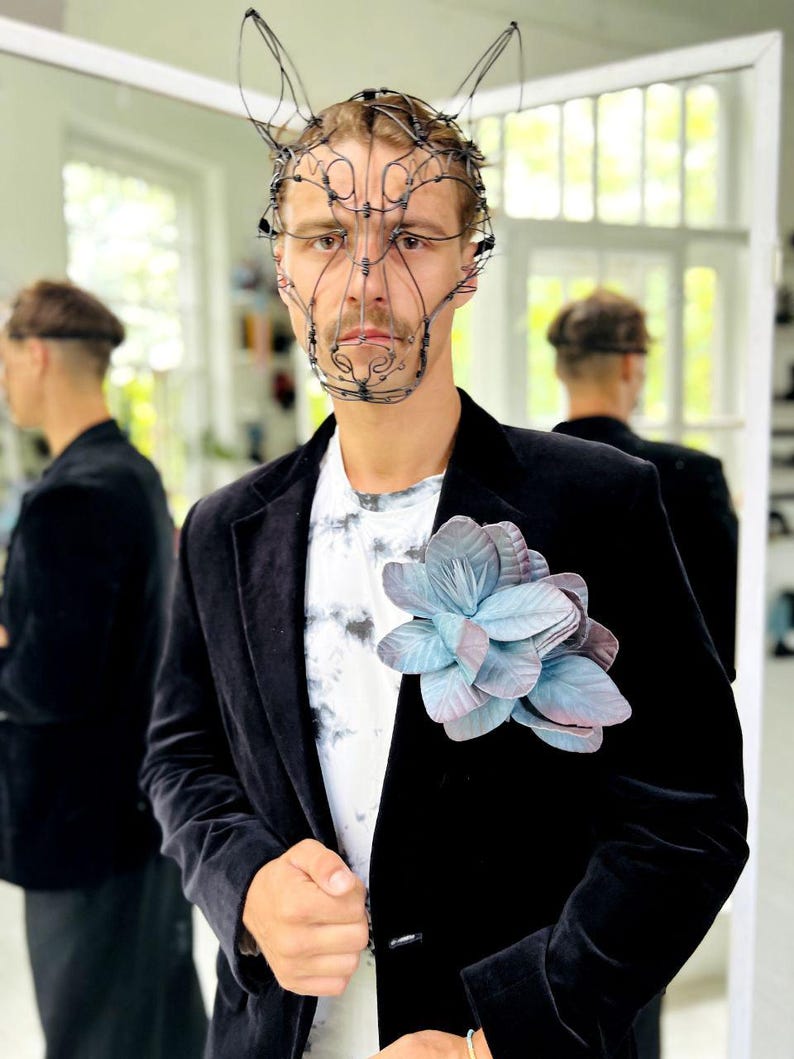 May include: A person wearing a black velvet blazer over a tie-dye t-shirt, adorned with a large, blue and purple flower. A wire sculpture mask with bunny ears is worn on the head.