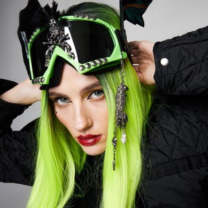 Puede incluir: Una persona con gafas de esquí verde neón con lentes negras, adornadas con adornos plateados y un lazo negro. La persona tiene el pelo verde brillante y lleva una chaqueta negra. Una cadena plateada con dijes cuelga de las gafas.