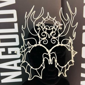 May include: A white wire crown with an intricate design featuring antler-like shapes and webbed patterns. The crown is displayed on a black mannequin head against a black background with white text.